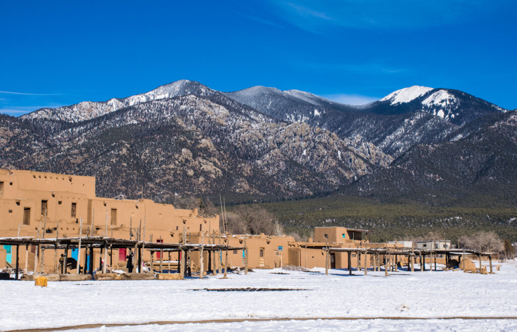 The Taos Pueblo on a sunny day with snow-capped mountains in the near background. 